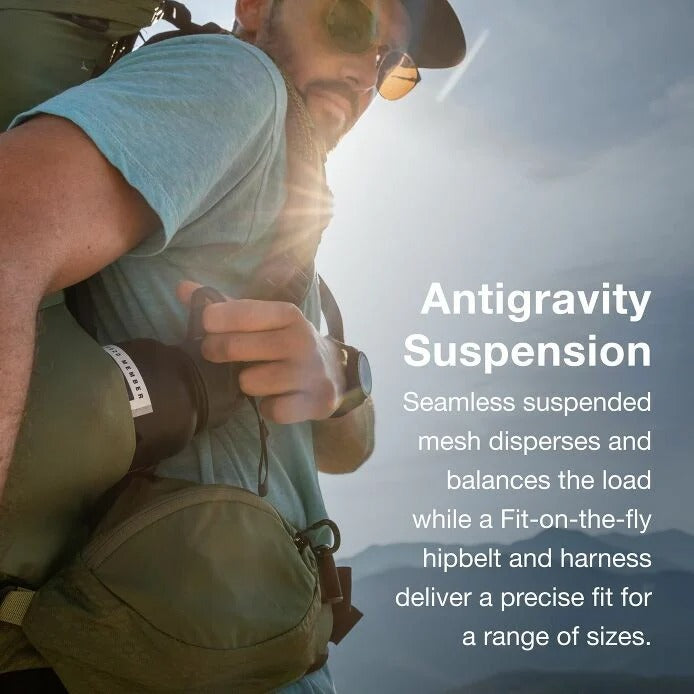 Person hiking with a backpack, wearing sunglasses and a cap, against a mountainous background.