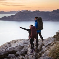 Two hikers with backpacks standing on a rocky outcrop overlooking a lake and mountains at sunset.