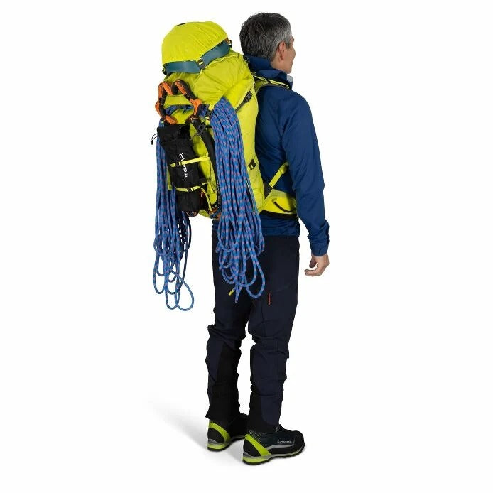 Person wearing a large yellow backpack with climbing equipment on a white background