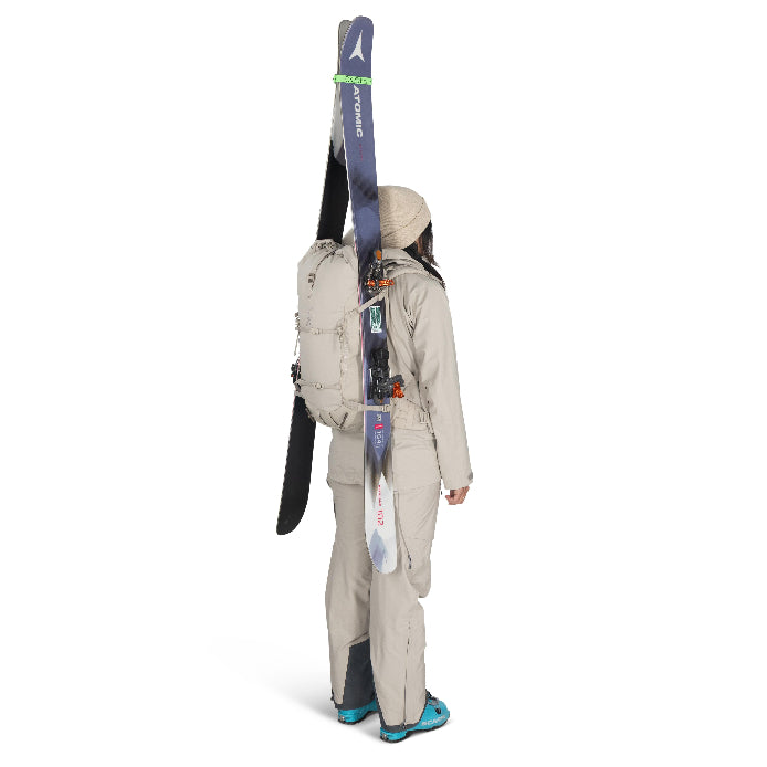 Person wearing a beige ski suit with skis attached to a backpack on a white background