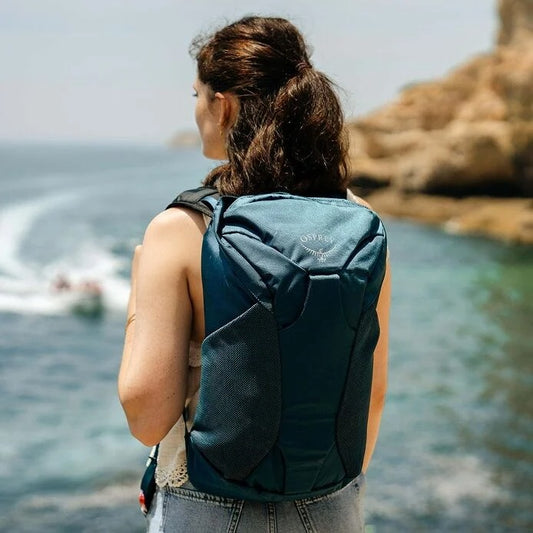 Person with a teal backpack standing by a body of water with a boat and rocky shore in the background.