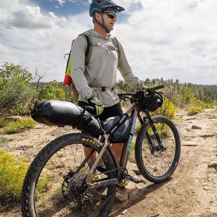 Man with a bicycle on a dirt trail with trees and sky in the background