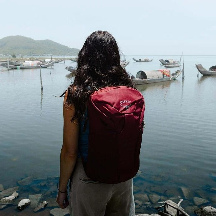 Person with a red Osprey backpack standing by a body of water with boats.
