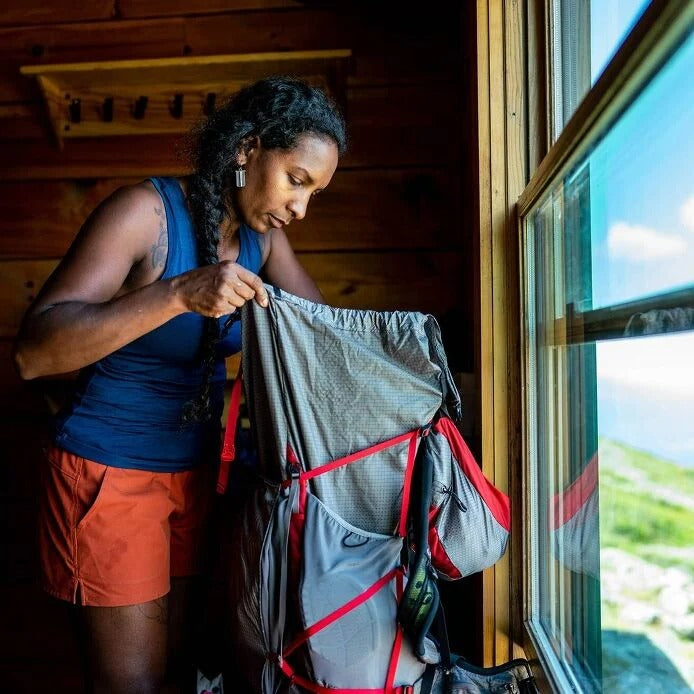 Person with a backpack inside a wooden cabin looking out a window with a scenic view.