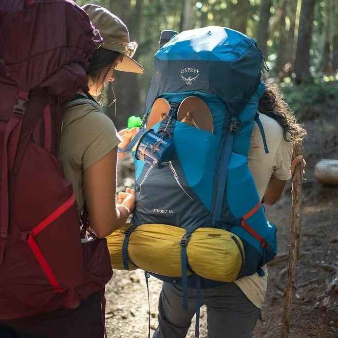 Two hikers with backpacks in a forest setting, one wearing a blue Osprey backpack.