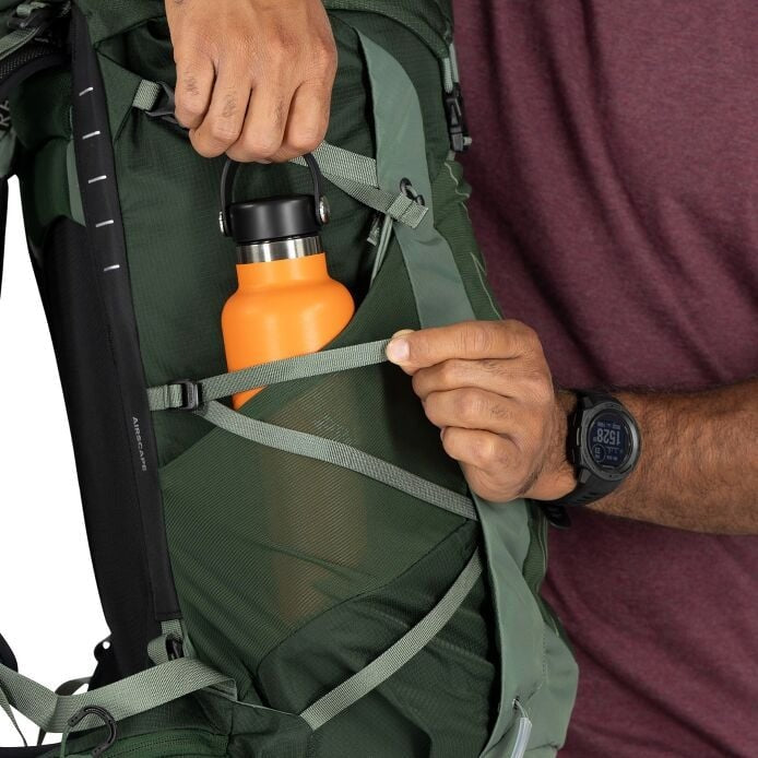 Person holding a green backpack with an orange water bottle and a rifle, wearing a purple shirt and black watch.