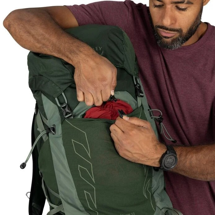 Man packing a green backpack with red clothing on a white background