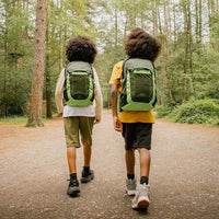 Two children walking on a path in a forest with green backpacks.