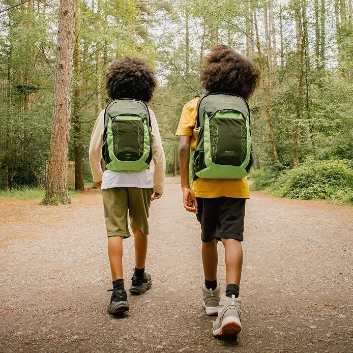 Two children walking on a path in a forest with green backpacks.